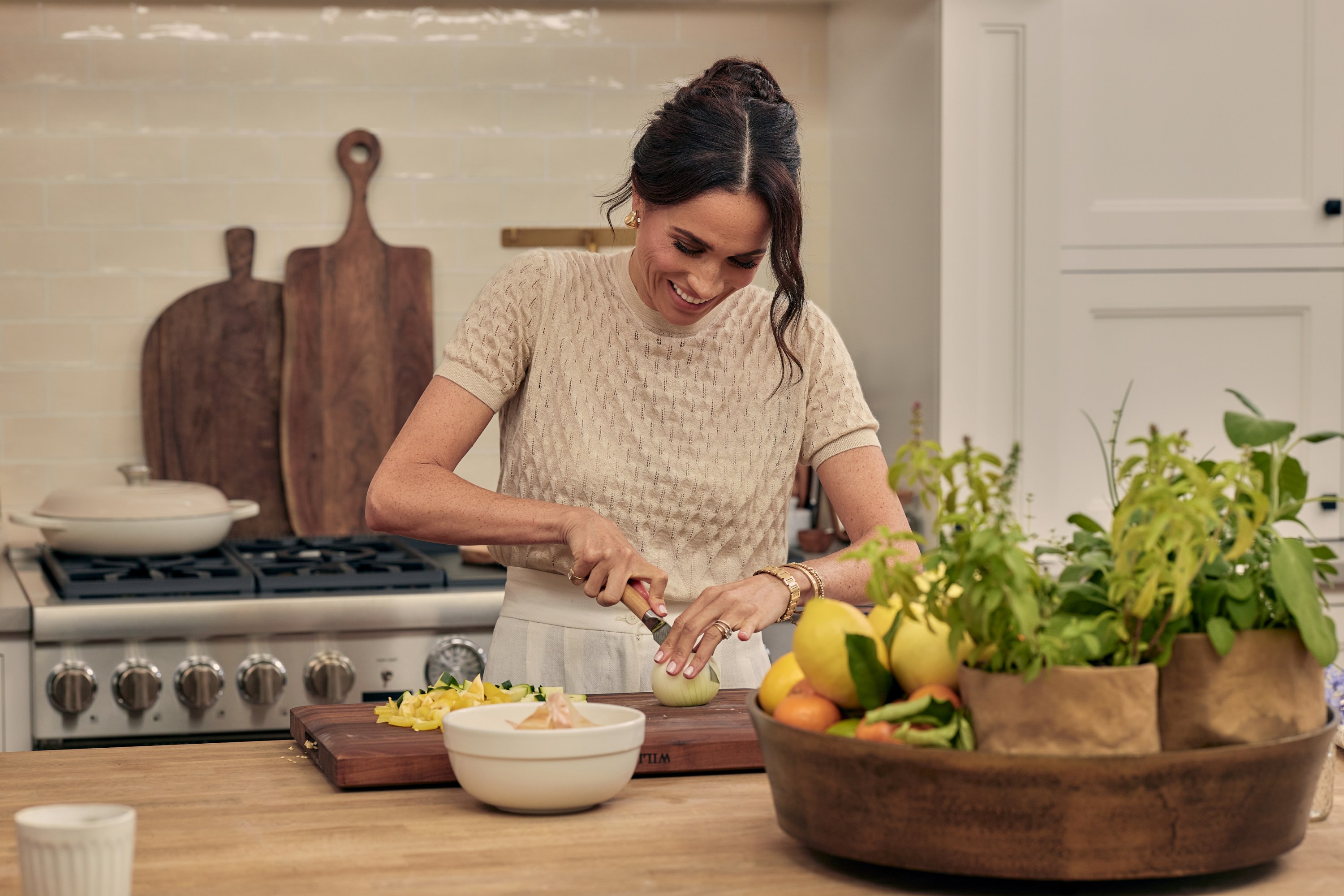 Meghan Markle in a kitchen, cutting an onion on a wooden cutting board.